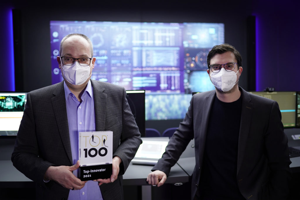 G&D CEOs Roland Ollek und Nils Strauch mit TOP-100-Award vor vielen Monitoren im ControlCenter-Xperience